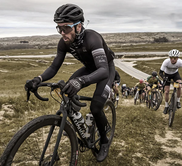 Un ciclista vestido de negro con casco y gafas lidera un grupo de competidores en una carrera de ciclismo de montaña o ruta por un sendero inclinado. Un ciclista vestido de negro con casco y gafas lidera un grupo de competidores en una carrera de ciclismo de montaña o ruta por un sendero inclinado.