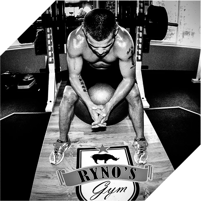 Un hombre atlético descansa sentado en el gimnasio con la cabeza baja, en una fotografía en blanco y negro que muestra el logo de RYNO'S Gym en el suelo.