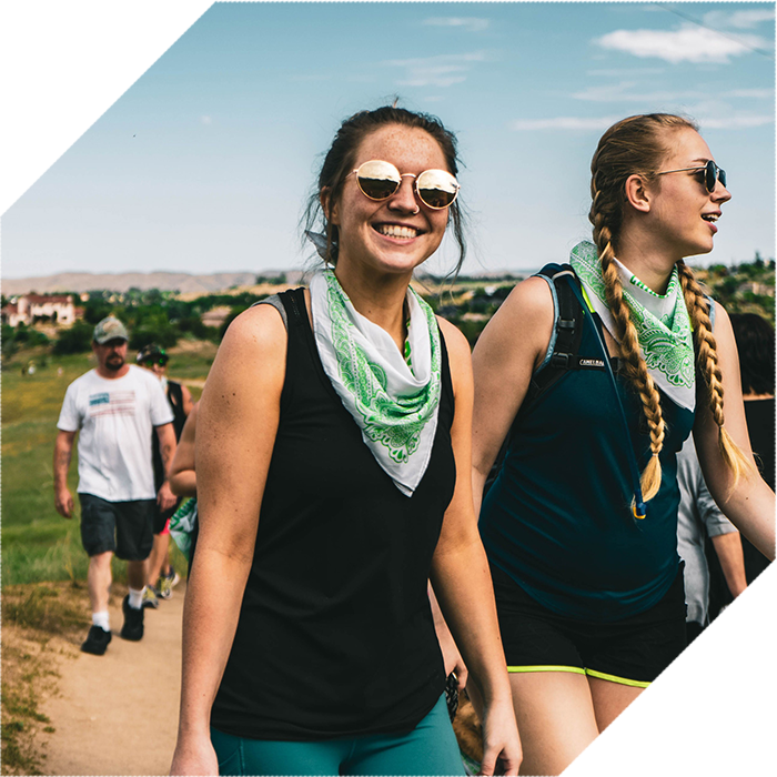 Dos jóvenes con gafas de sol y pañuelos verdes al cuello caminan sonrientes por un sendero al aire libre junto a otros excursionistas.