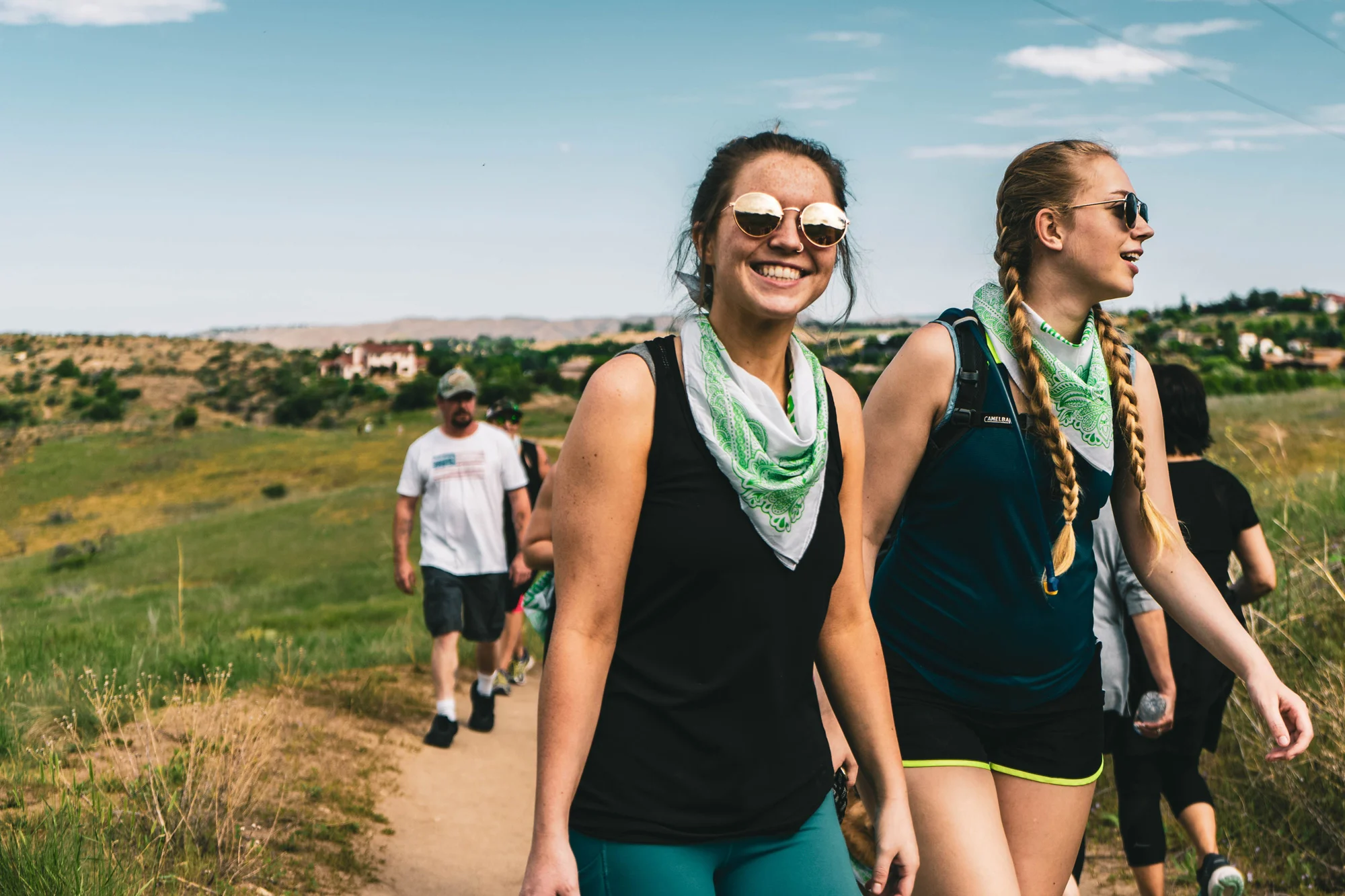Dos jóvenes con gafas de sol y pañuelos verdes al cuello caminan sonrientes por un sendero al aire libre junto a otros excursionistas.
