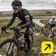 Un ciclista vestido de negro con casco y gafas lidera un grupo de competidores en una carrera de ciclismo de montaña o ruta por un sendero inclinado.